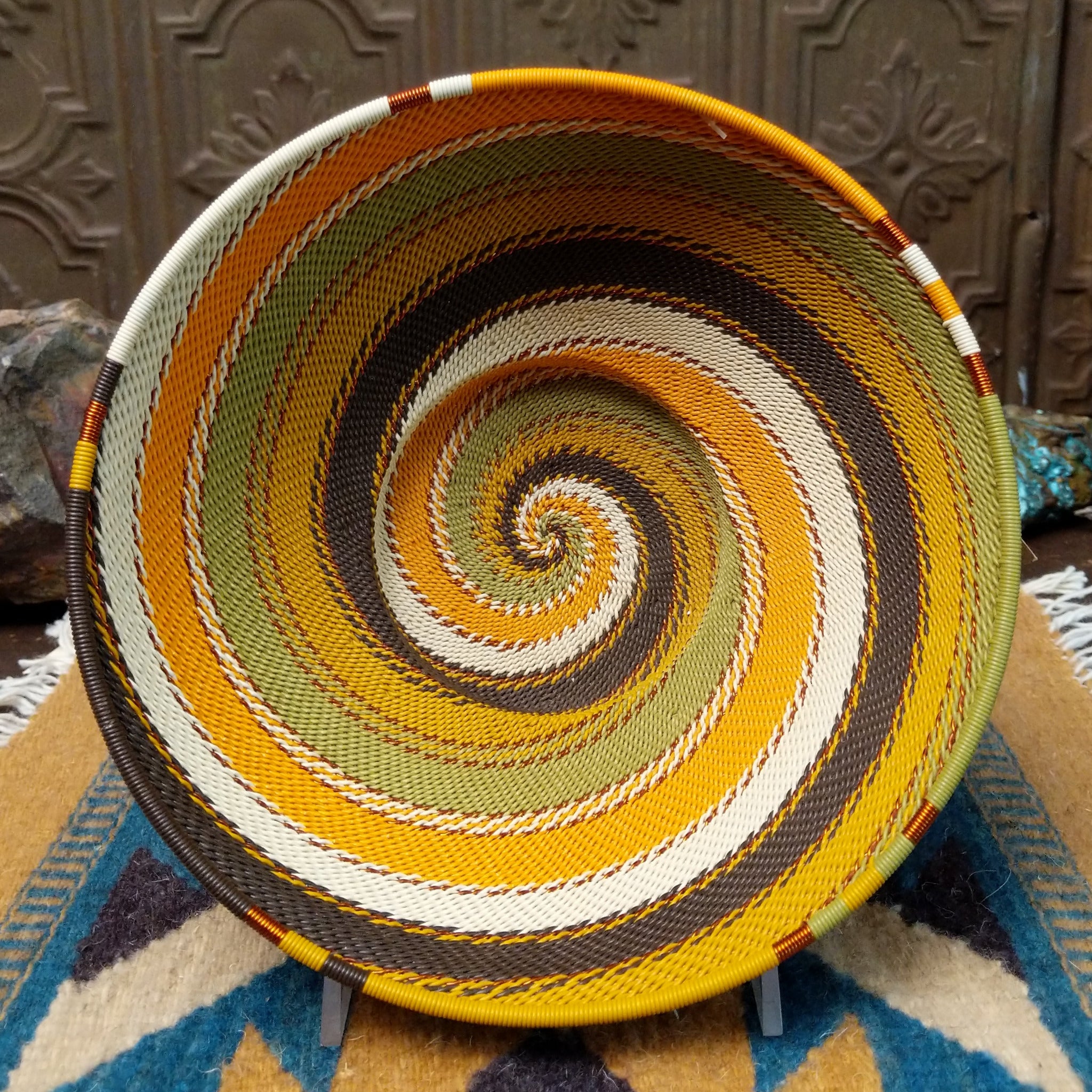 Handwoven earth tone wire art bowl by Bridge for Africa featuring a spiral pattern in recycled wire with golden yellow, olive green, ivory, brown, and tan tones, perfect for home décor or wall display.