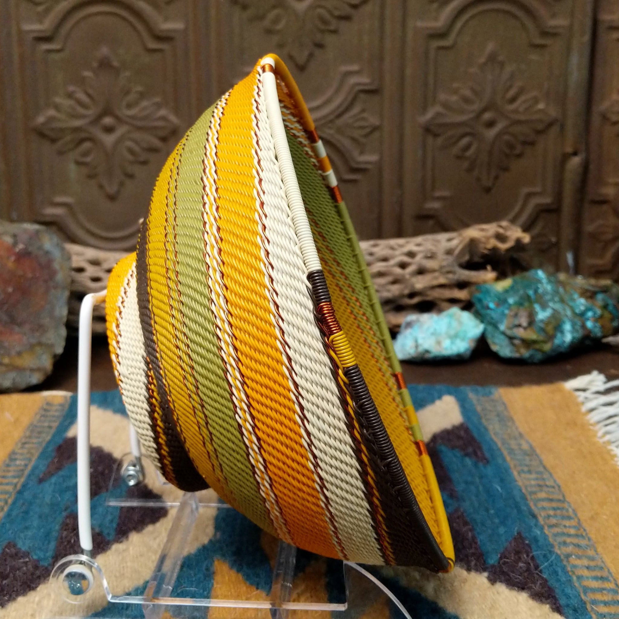 Handwoven earth tone wire art bowl by Bridge for Africa featuring a spiral pattern in recycled wire with golden yellow, olive green, ivory, brown, and tan tones, perfect for home décor or wall display; side view.