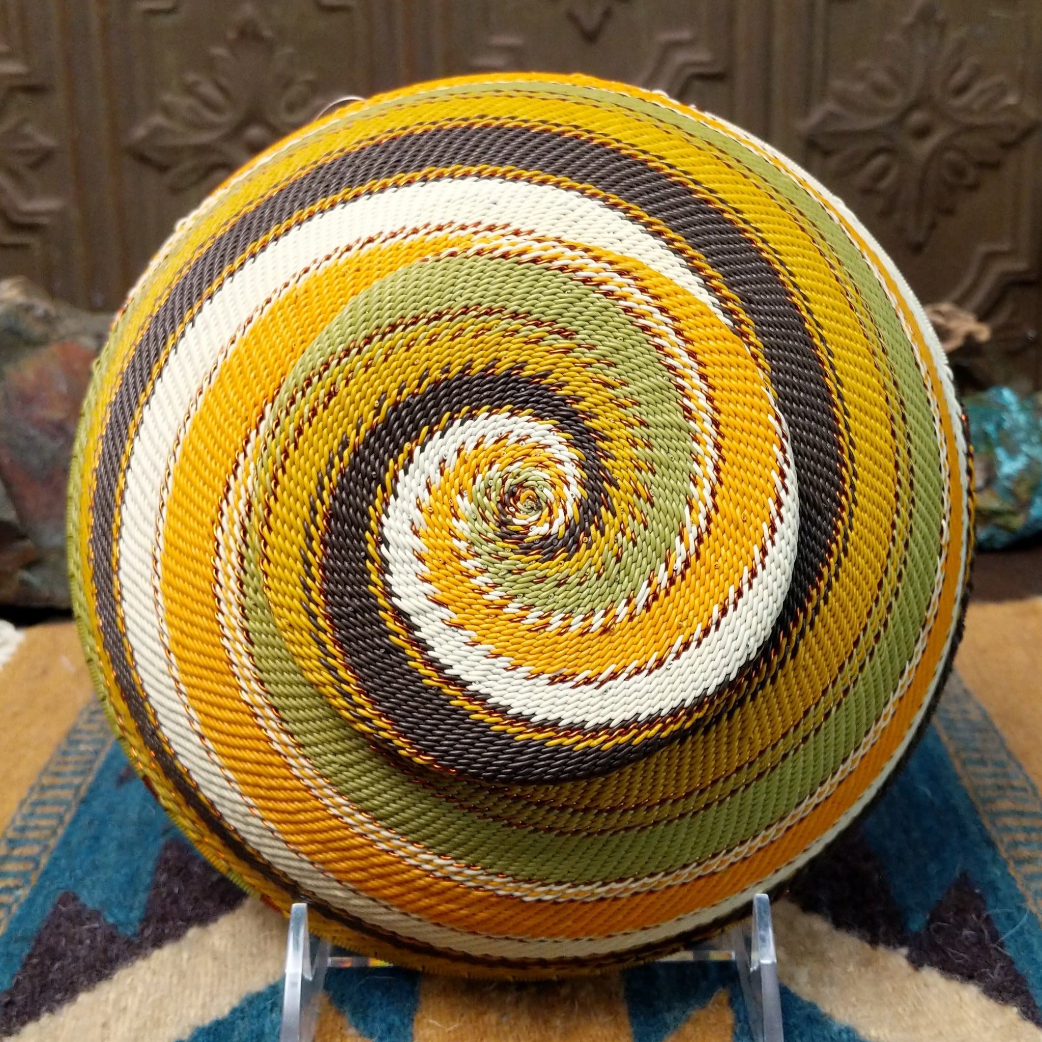 Handwoven earth tone wire art bowl by Bridge for Africa featuring a spiral pattern in recycled wire with golden yellow, olive green, ivory, brown, and tan tones, perfect for home décor or wall display; underside view.