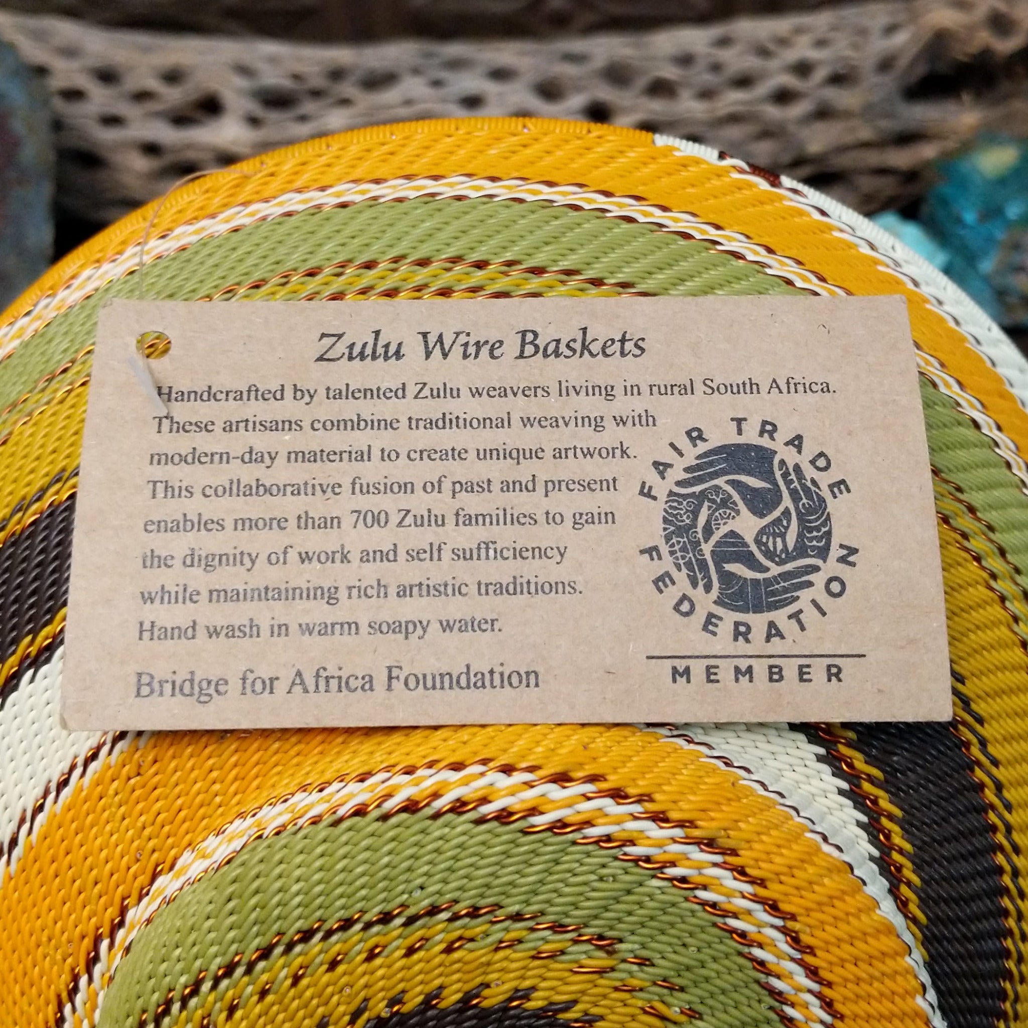 Handwoven earth tone wire art bowl by Bridge for Africa featuring a spiral pattern in recycled wire with golden yellow, olive green, ivory, brown, and tan tones, perfect for home décor or wall display; artisan information.