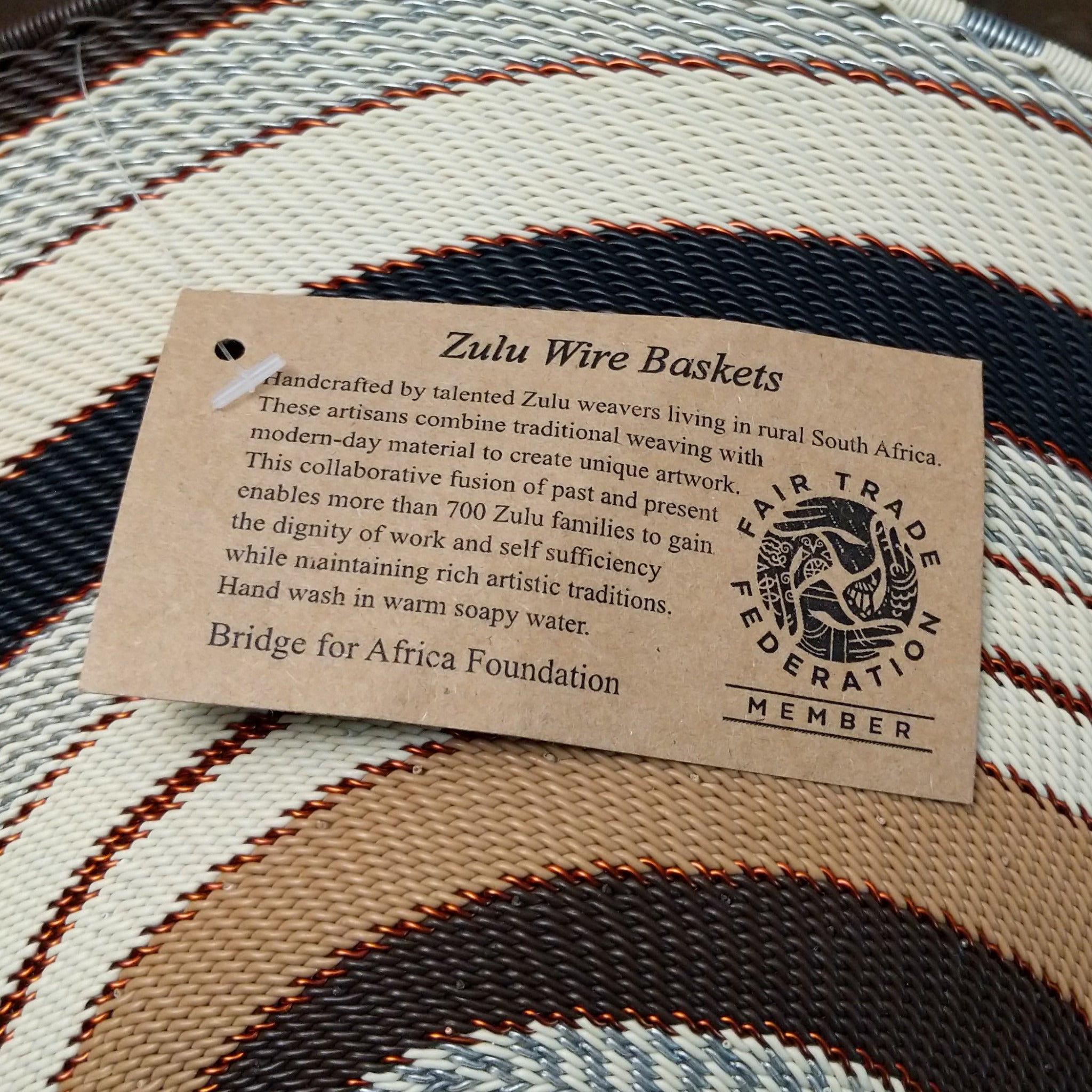 Handwoven large earth tone wire art bowl by Bridge for Africa featuring a spiral pattern in recycled wire with ivory, beige, taupe, gray, brown, and black tones, perfect for home décor or wall display; artisan information.