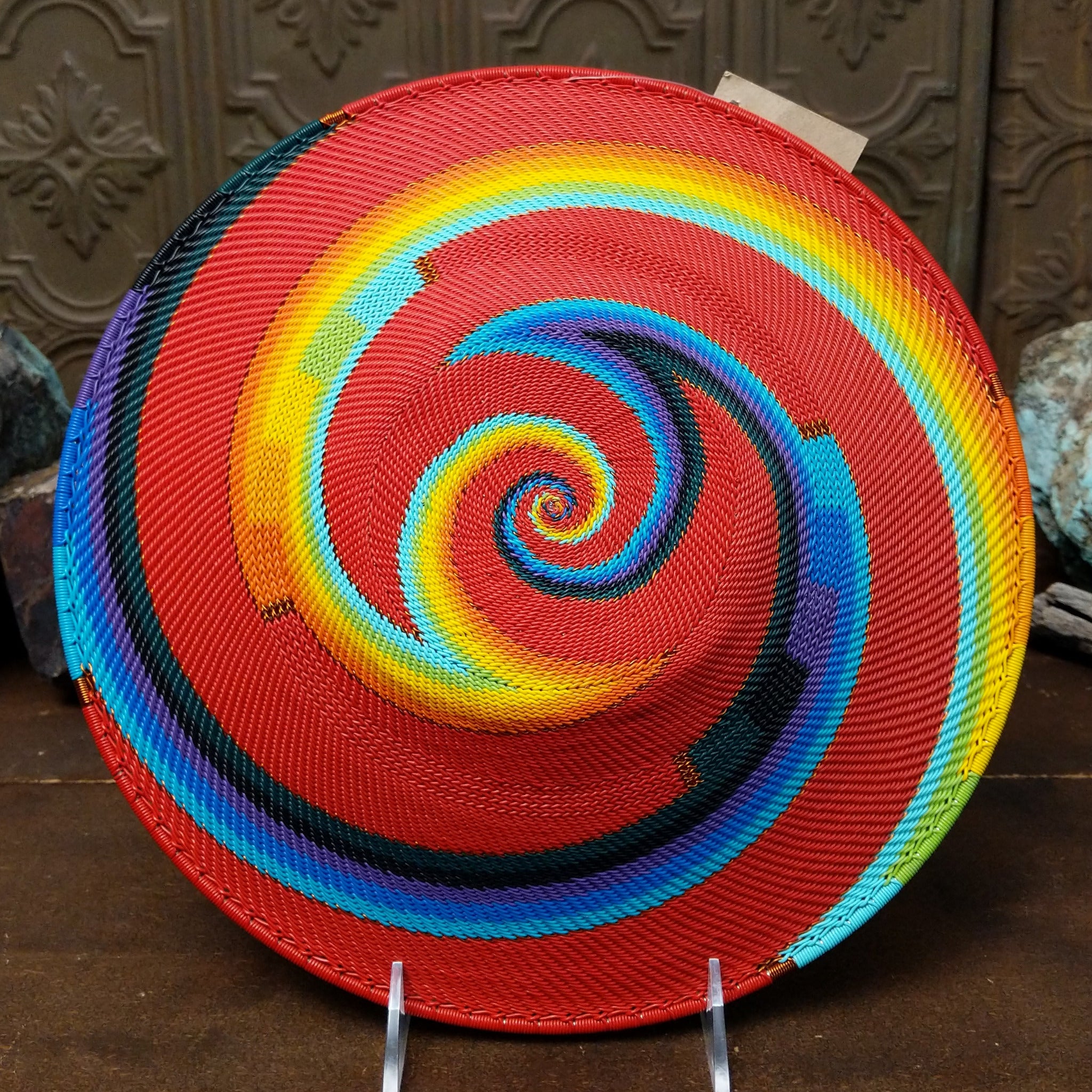 Handwoven large multicolor wire art basket by Bridge for Africa featuring a vibrant spiral pattern in recycled wire with bold red, blue, turquoise, green, yellow, orange, purple, and black accents; underside view.