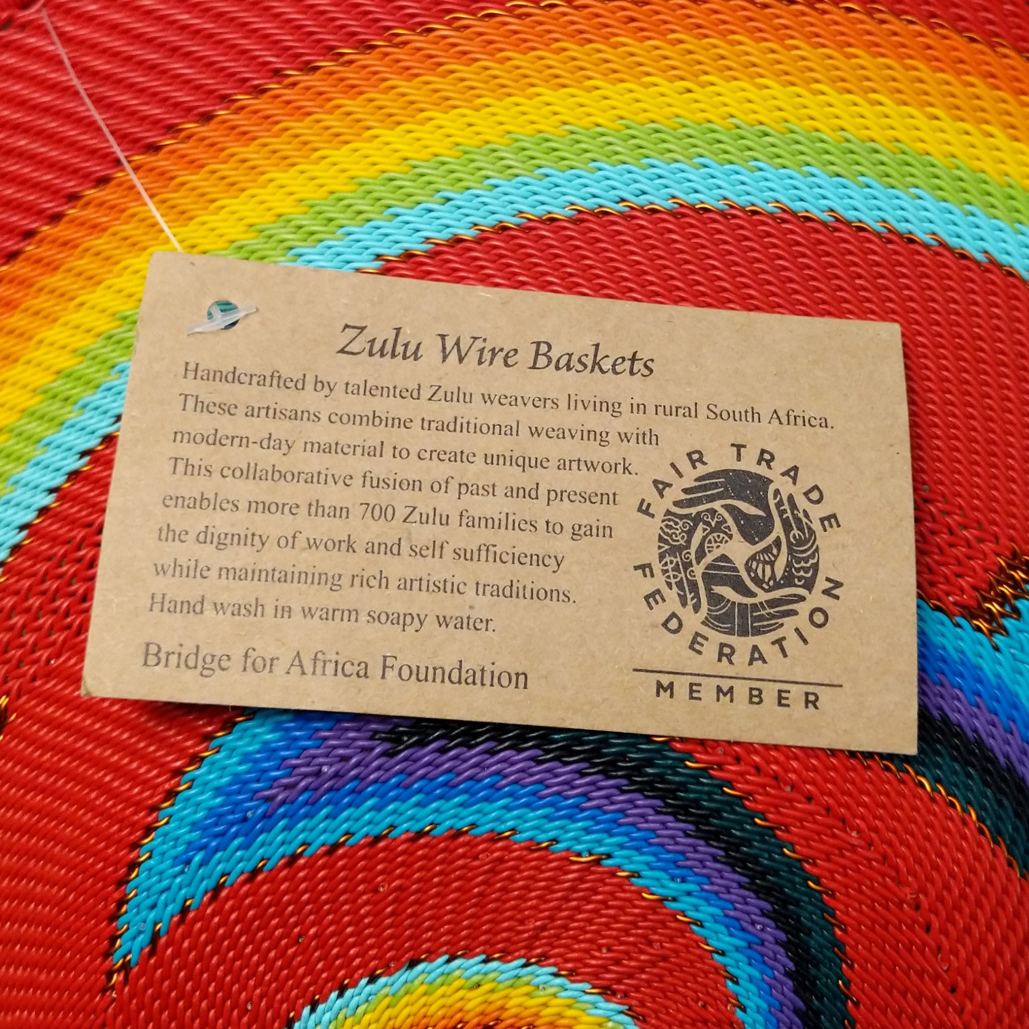 Handwoven large multicolor wire art basket by Bridge for Africa featuring a vibrant spiral pattern in recycled wire with bold red, blue, turquoise, green, yellow, orange, purple, and black accents; artisan information.