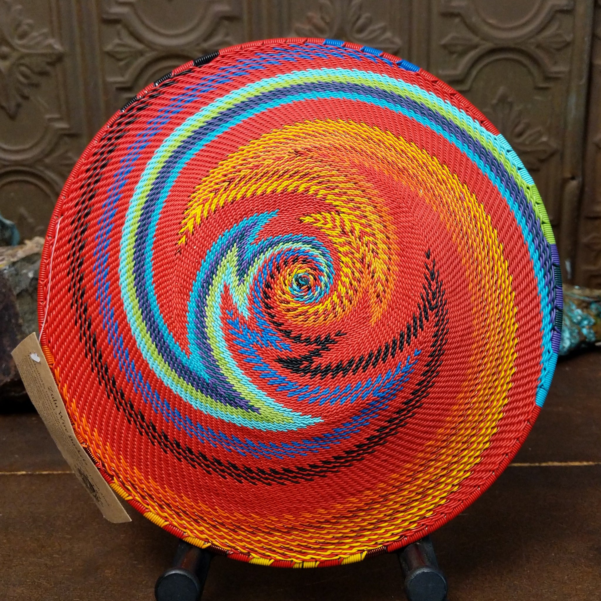 Handwoven multicolor wire art basket by Bridge for Africa featuring a vibrant spiral pattern made from recycled wire in bold rainbow colors, ideal for display or home décor; underside view.