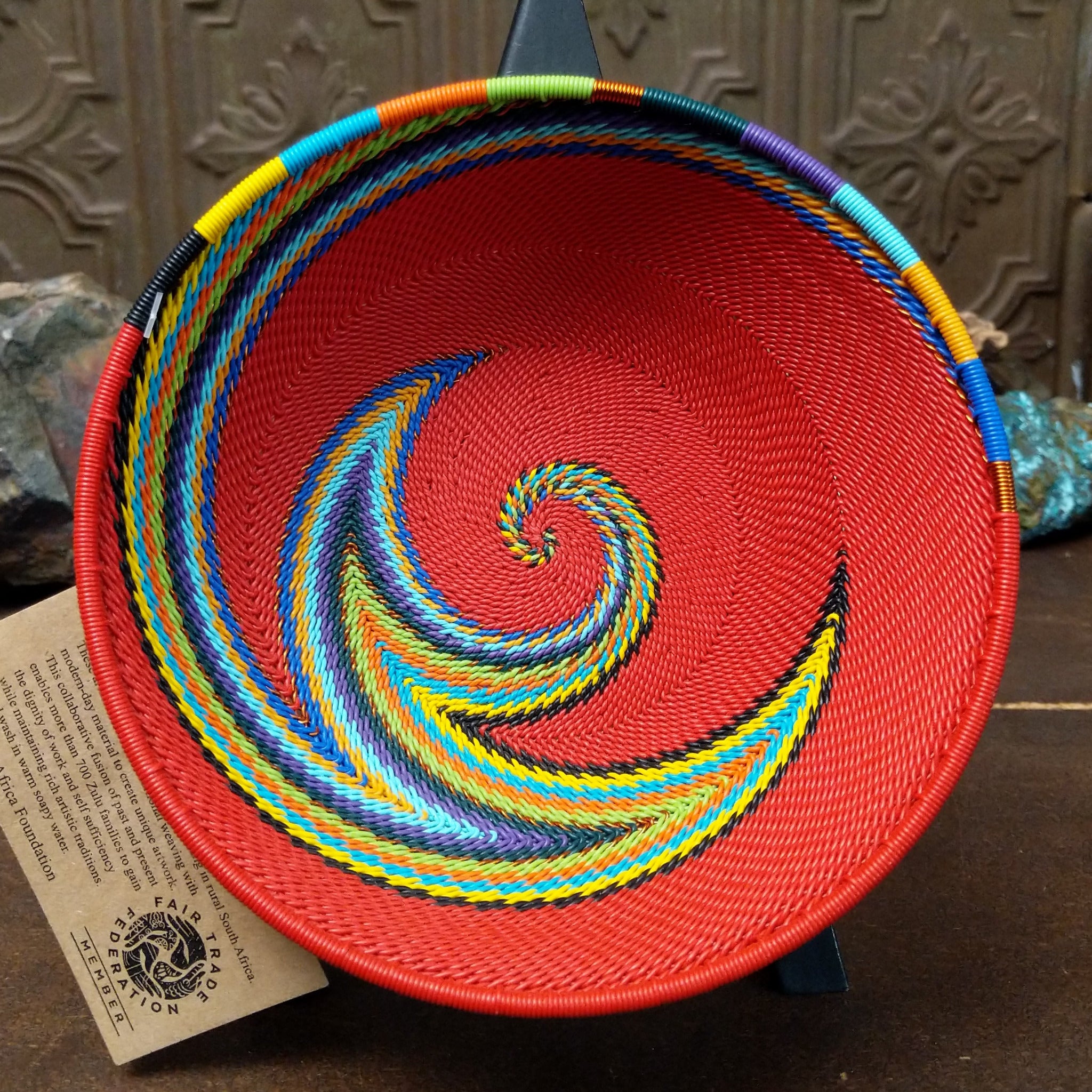 Handwoven multicolor wire art bowl by Bridge for Africa featuring a vibrant spiral design made from recycled wire in bold red, blue, green, yellow, orange, purple, and black tones.