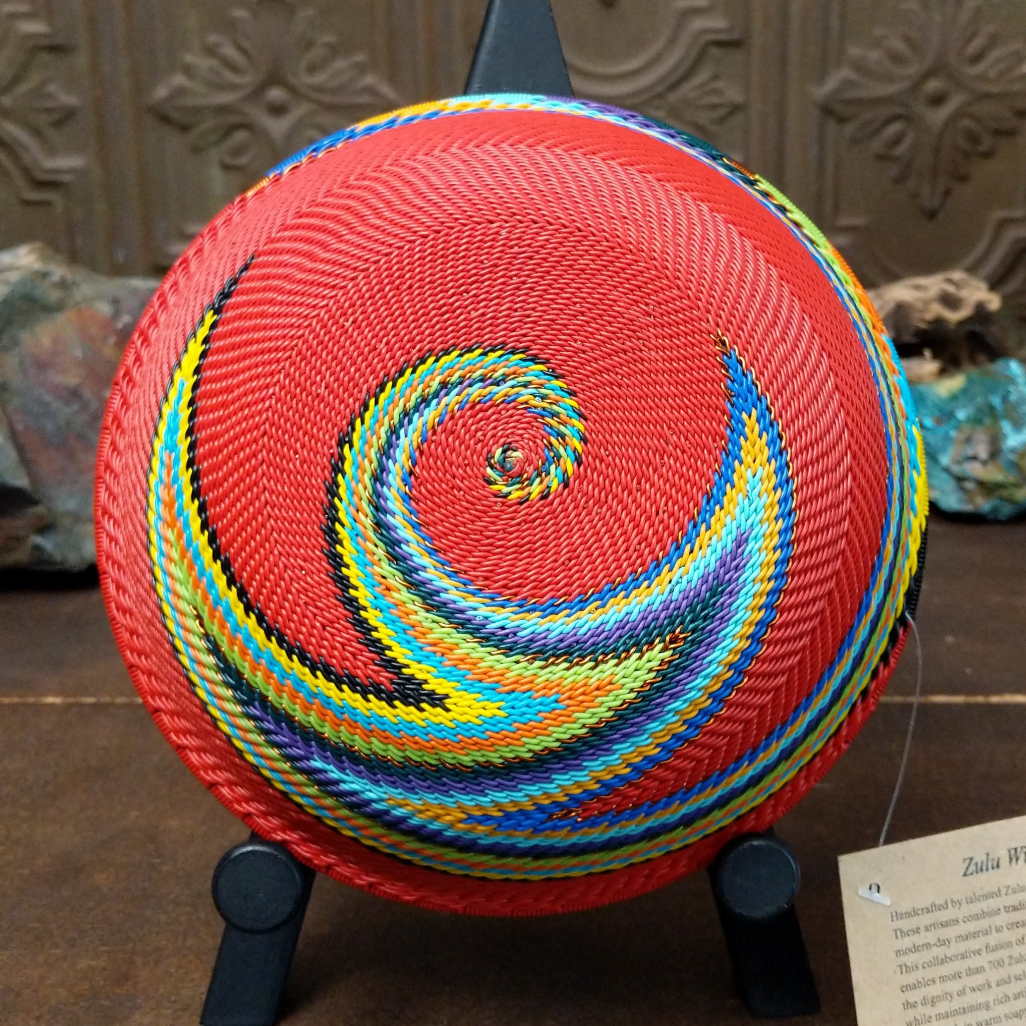 Handwoven multicolor wire art bowl by Bridge for Africa featuring a vibrant spiral design made from recycled wire in bold red, blue, green, yellow, orange, purple, and black tones; underside view.