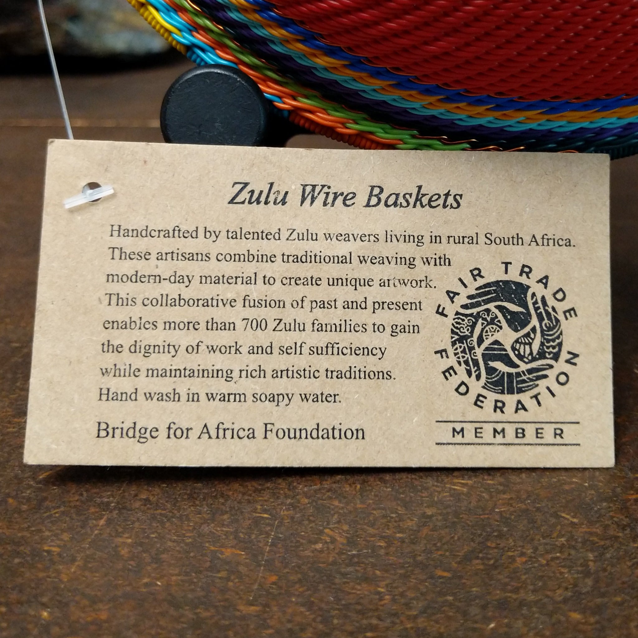 Handwoven multicolor wire art bowl by Bridge for Africa featuring a vibrant spiral design made from recycled wire in bold red, blue, green, yellow, orange, purple, and black tones; artisan information.