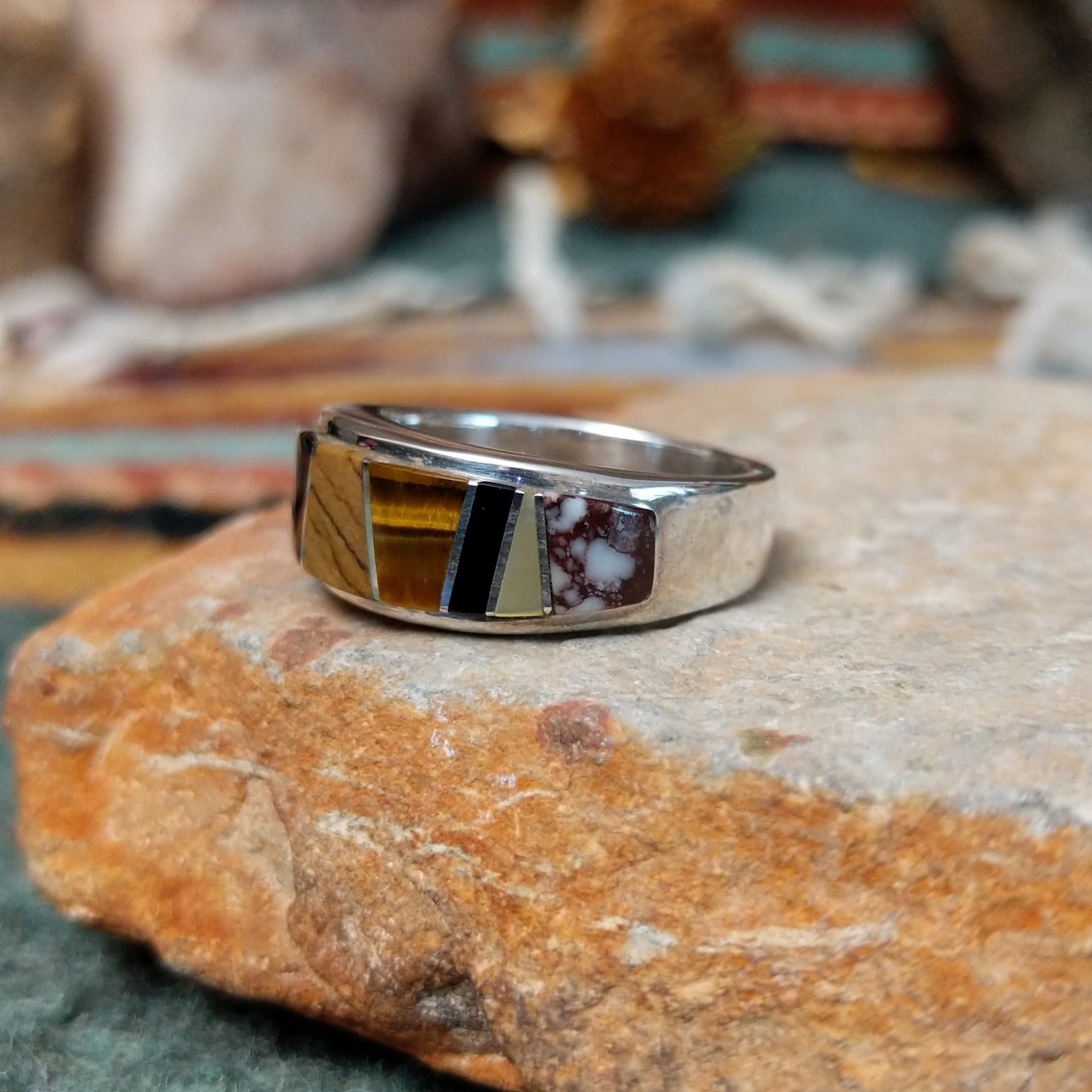 Navajo sterling silver ring by J.C. featuring geometric multistone inlay with black onyx, tiger’s eye, and assorted natural stones separated by silver dividers; side view.