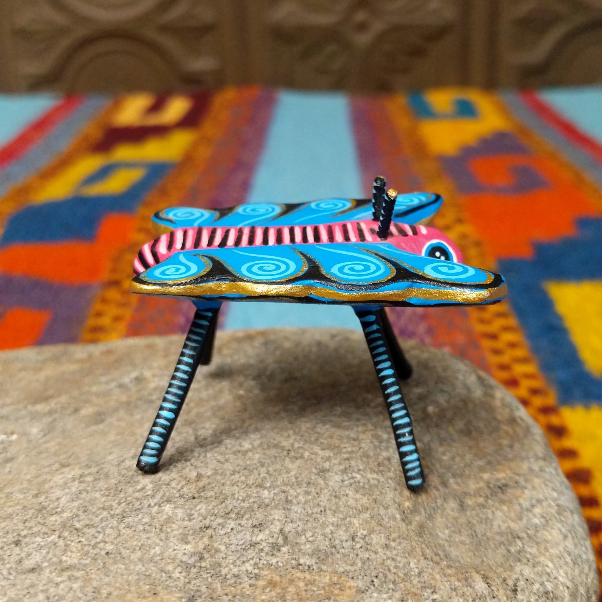 This colorful Oaxacan butterfly wood carving features a pink body, turquoise wings with spiral patterns, gold accents, striped antennae, and long black-and-turquoise legs; side view.