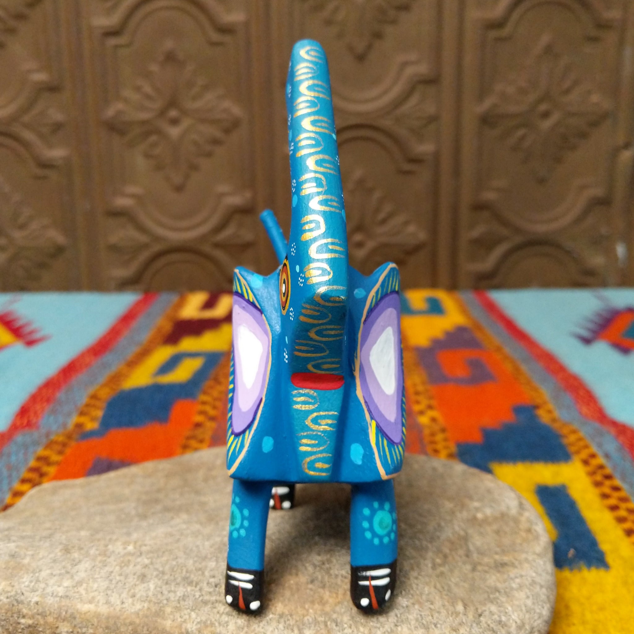 This blue Oaxacan elephant wood carving by Roberta Angeles features an uplifted trunk, lavender ear, pink flower, and colorful hand-painted decorative patterns; front view.