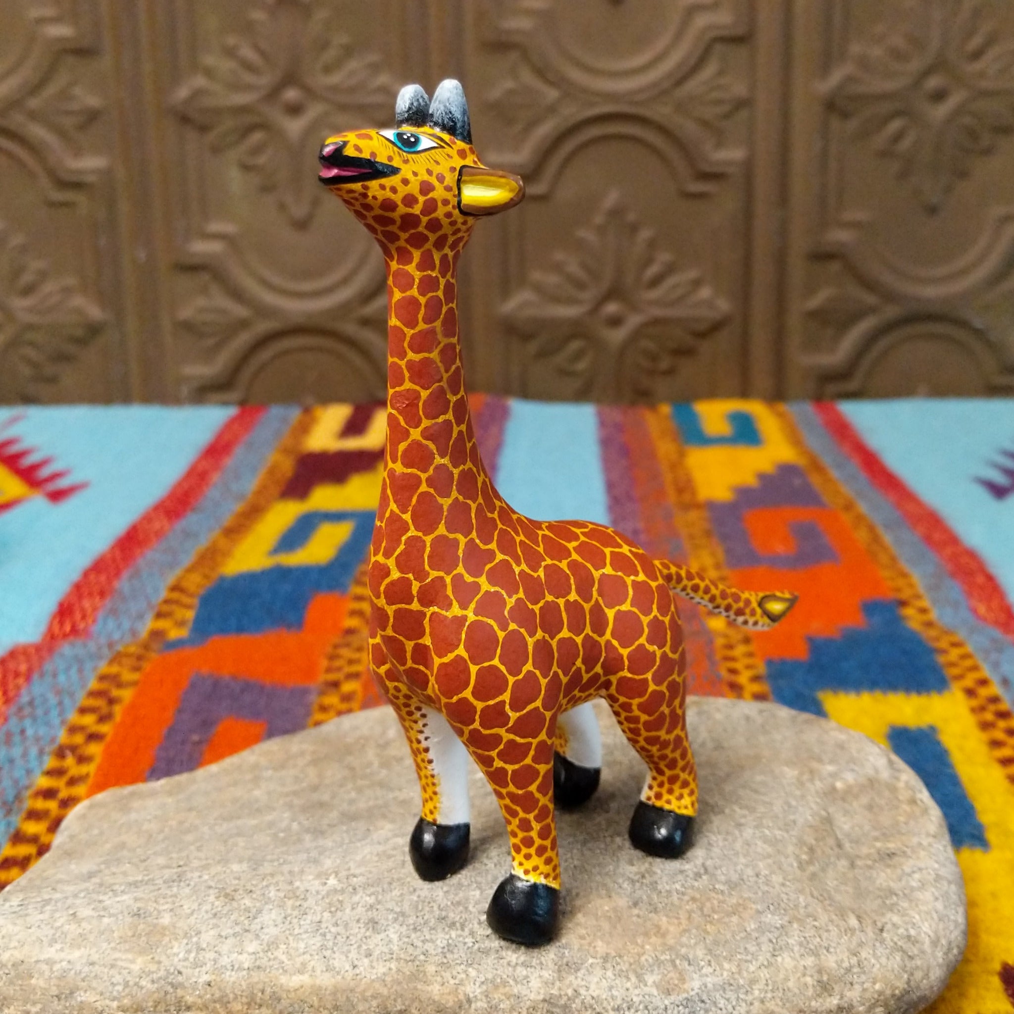 This Oaxacan giraffe wood carving by Faviola Fabian features a golden-yellow body with reddish-brown spots, a long neck, blue eyes, black-and-white horns, and black hooves.