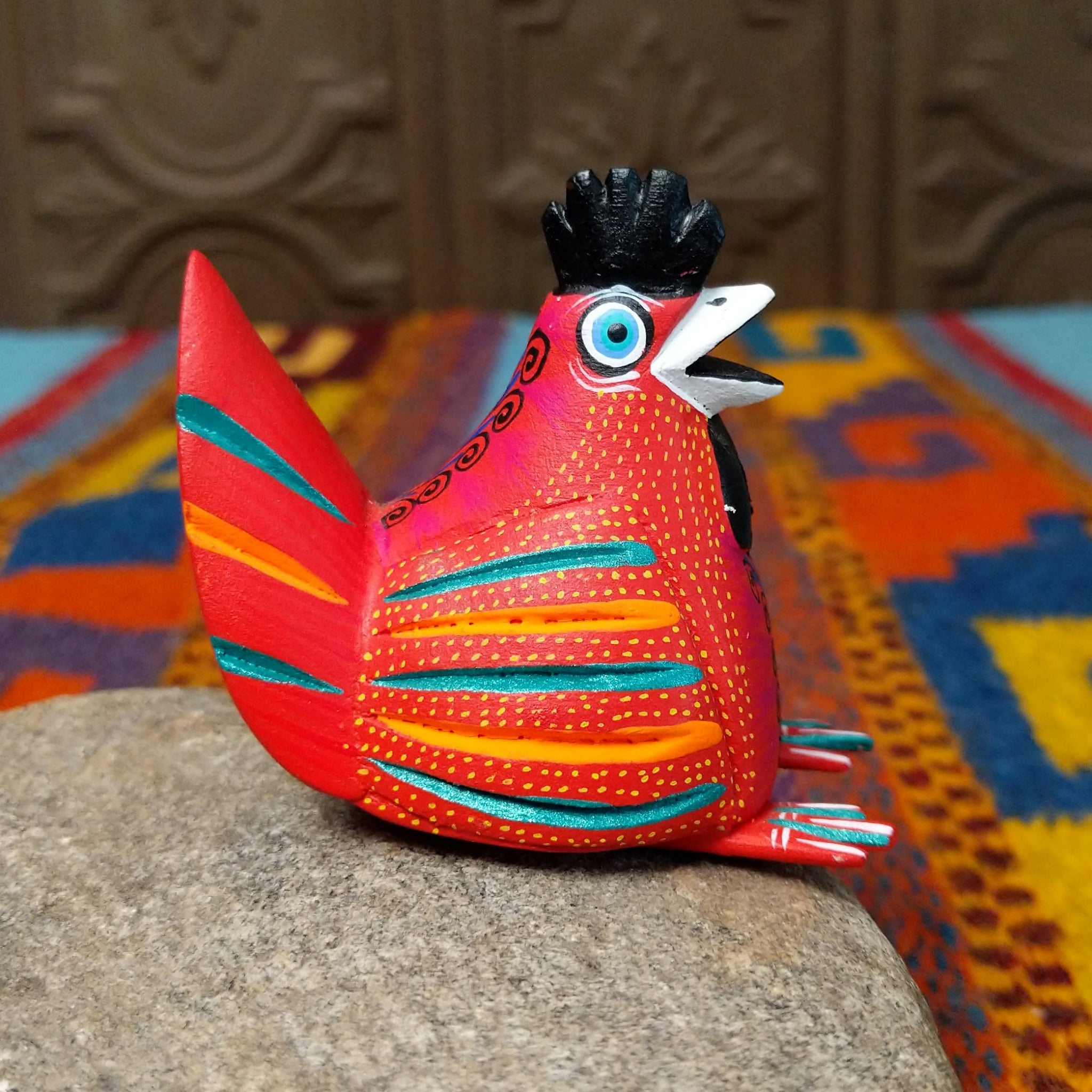 This colorful Oaxacan hen wood carving features a bright red body, black comb, turquoise eye, and intricate yellow, orange, turquoise, and black hand-painted feather patterns; side view.