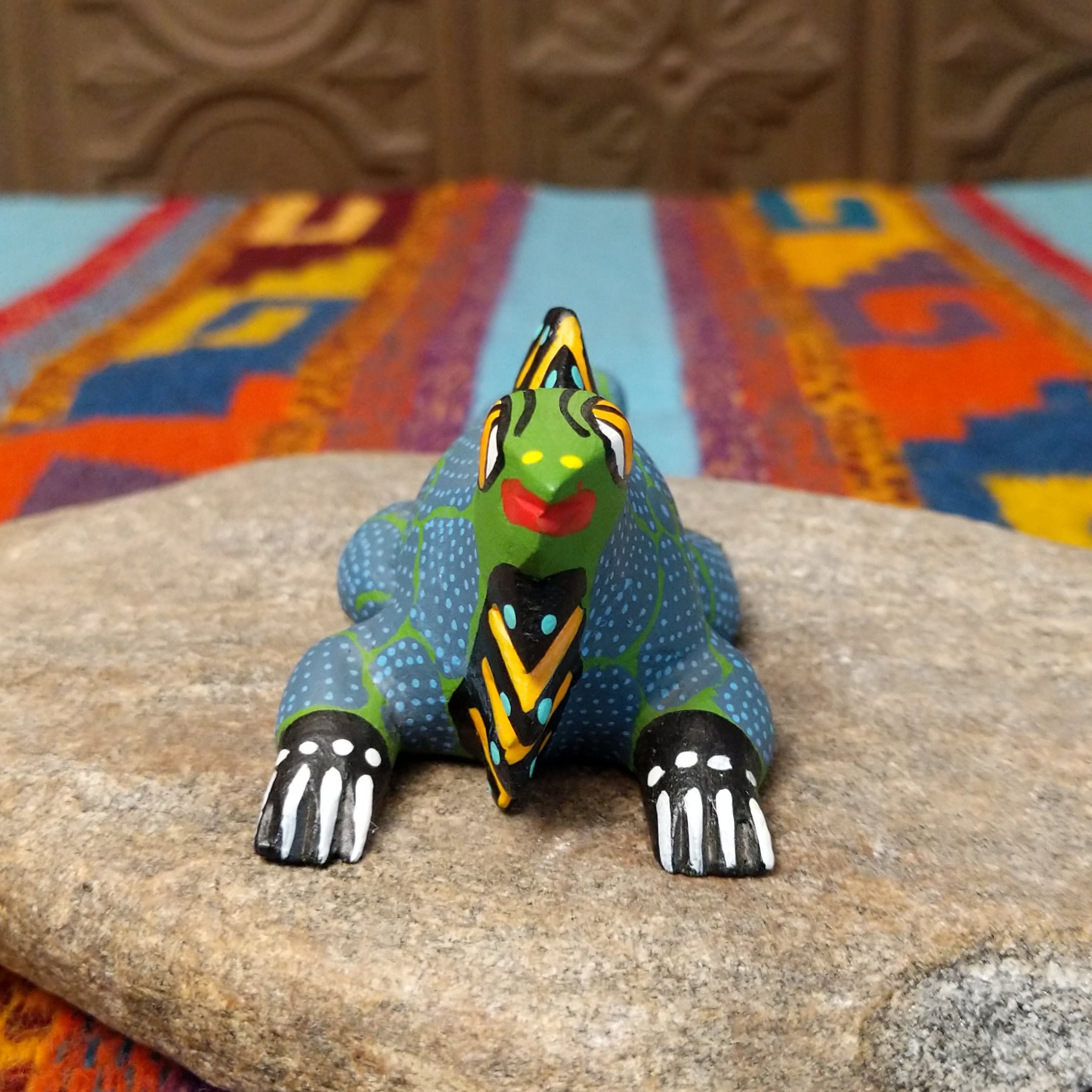 This colorful Oaxacan iguana wood carving features a green head, blue dotted body, golden eyes, black-and-yellow back spines, and a long patterned tail; front facing view.