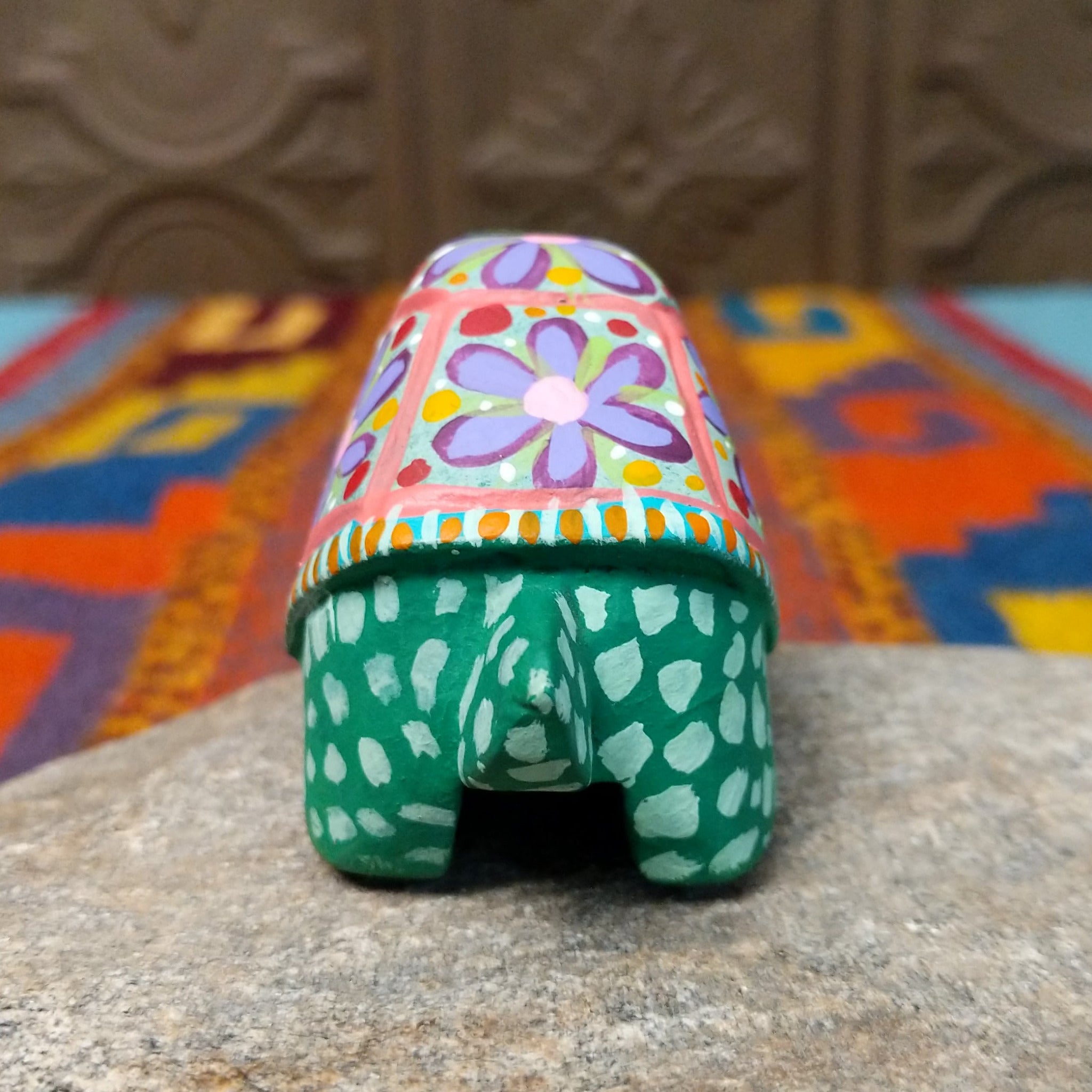 This green Oaxacan tortoise wood carving by Rosaura Ortega features a domed shell decorated with purple flowers, pink borders, and colorful hand-painted dots and patterns; rear view.