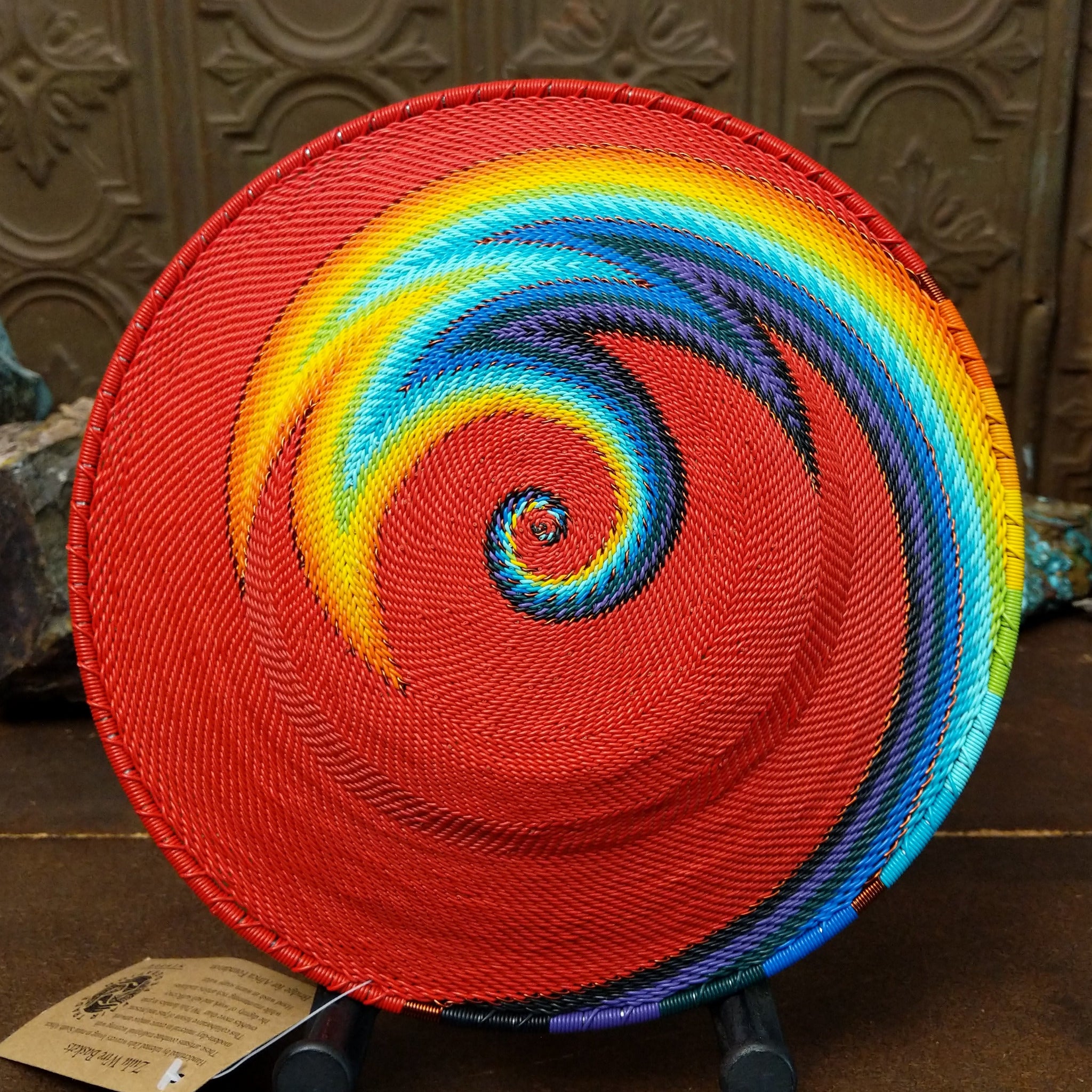 Handwoven rainbow wire art basket by Bridge for Africa featuring colorful recycled wire in a vibrant geometric pattern, ideal as a decorative bowl or wall display; underside view.