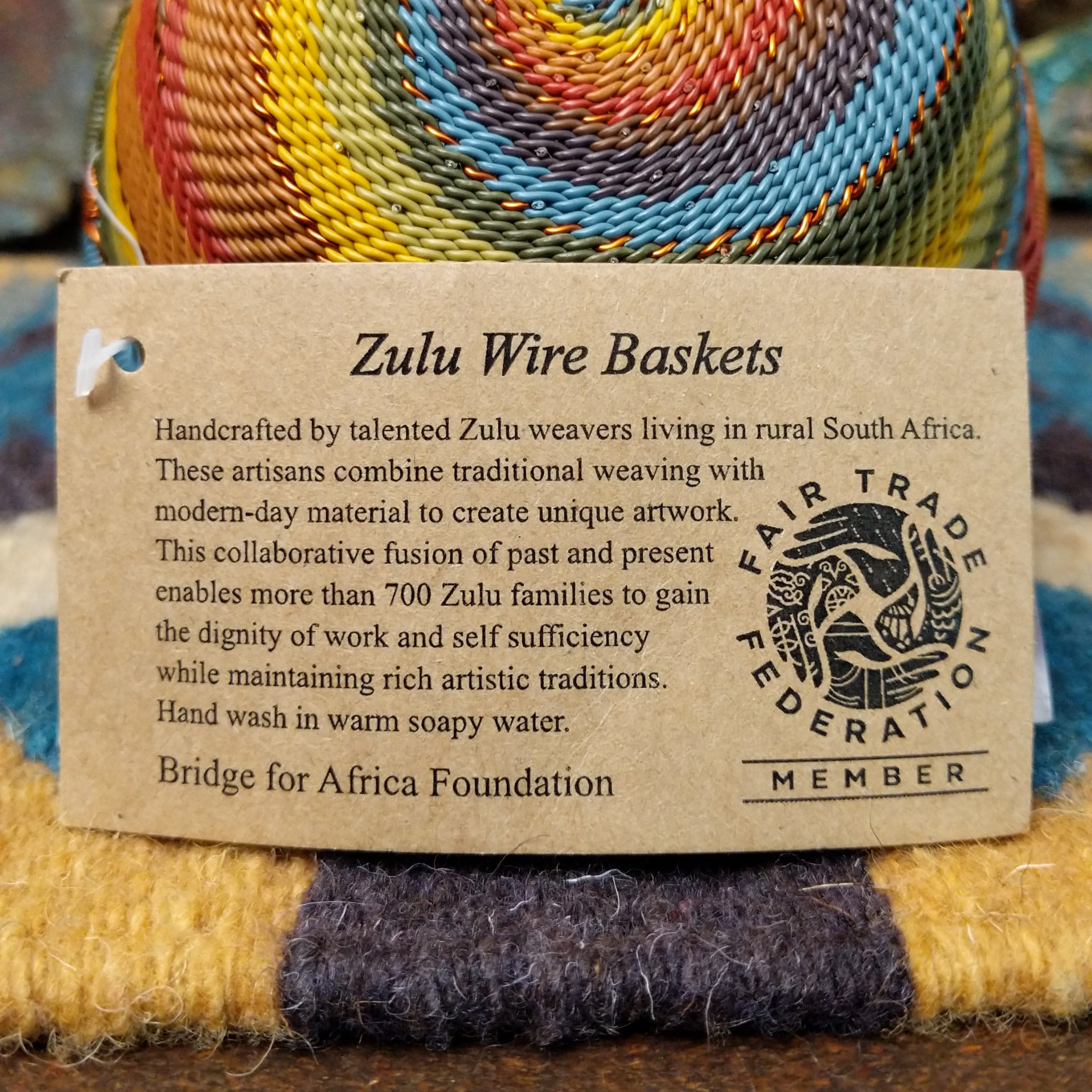 Handwoven small wire art bowl by Bridge for Africa featuring a spiral pattern in recycled wire with mustard yellow, olive green, dusty blue, rust, terracotta, brown, and charcoal tones, ideal for home décor or as a decorative catchall; artisan information.