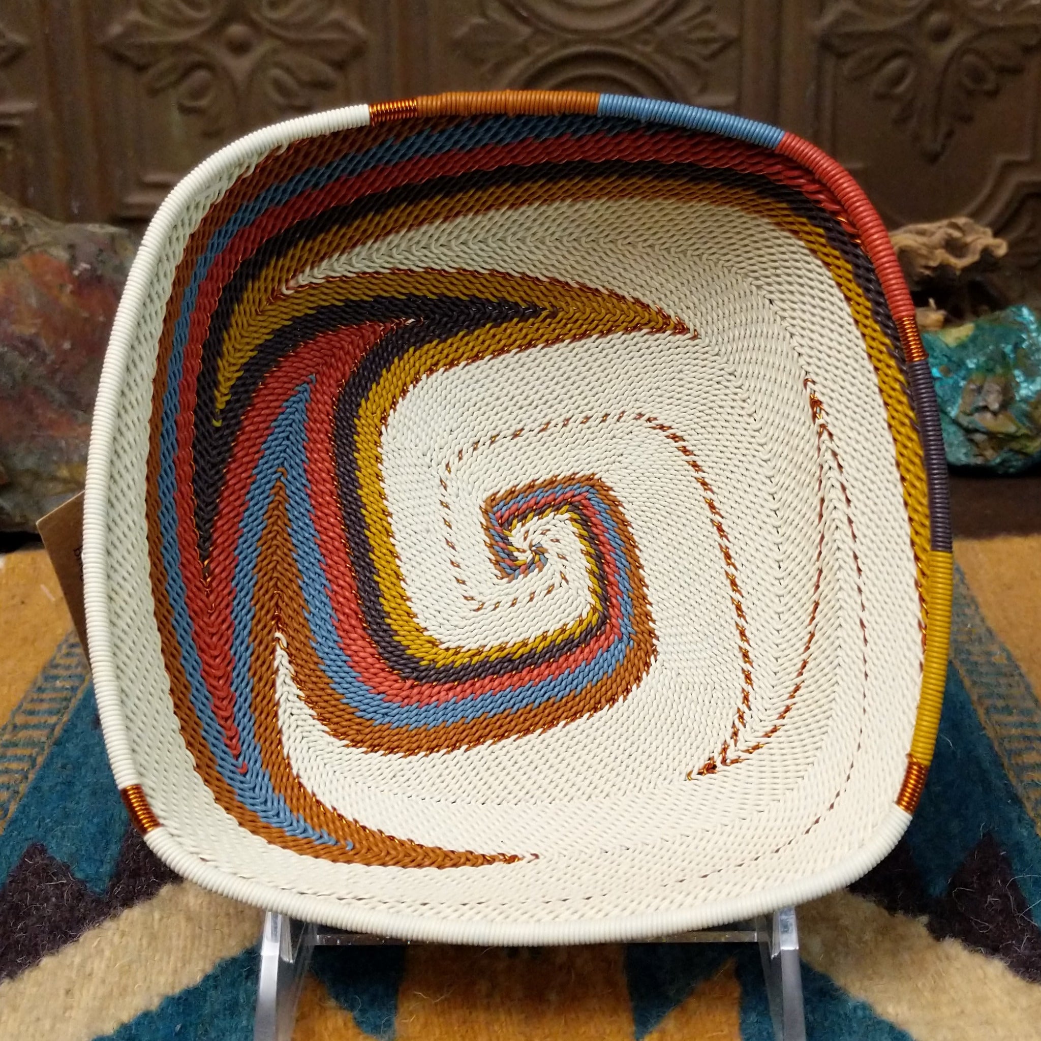 Handwoven square wire art bowl by Bridge for Africa featuring a spiral pattern in recycled wire with ivory, rust, blue, gold, brown, and charcoal tones, ideal for home décor or wall display.