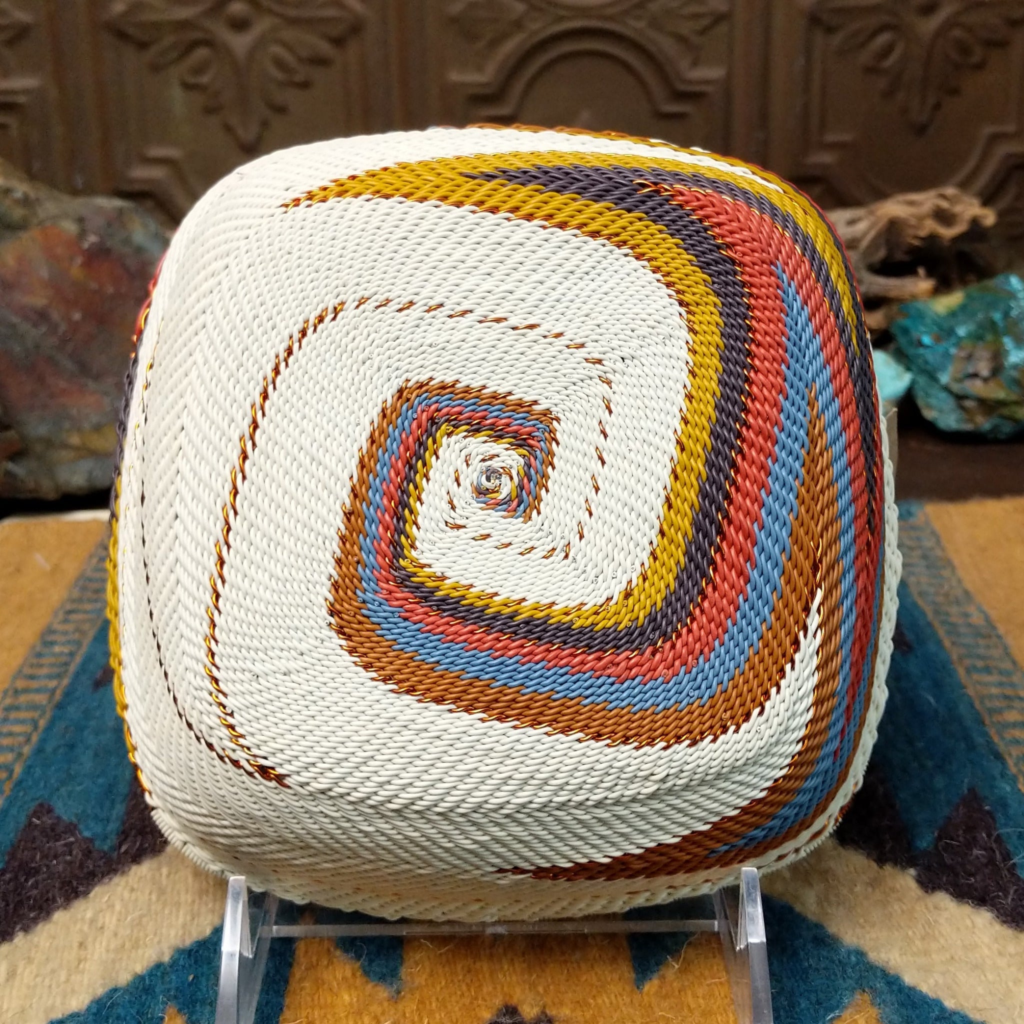 Handwoven square wire art bowl by Bridge for Africa featuring a spiral pattern in recycled wire with ivory, rust, blue, gold, brown, and charcoal tones, ideal for home décor or wall display; underside view.