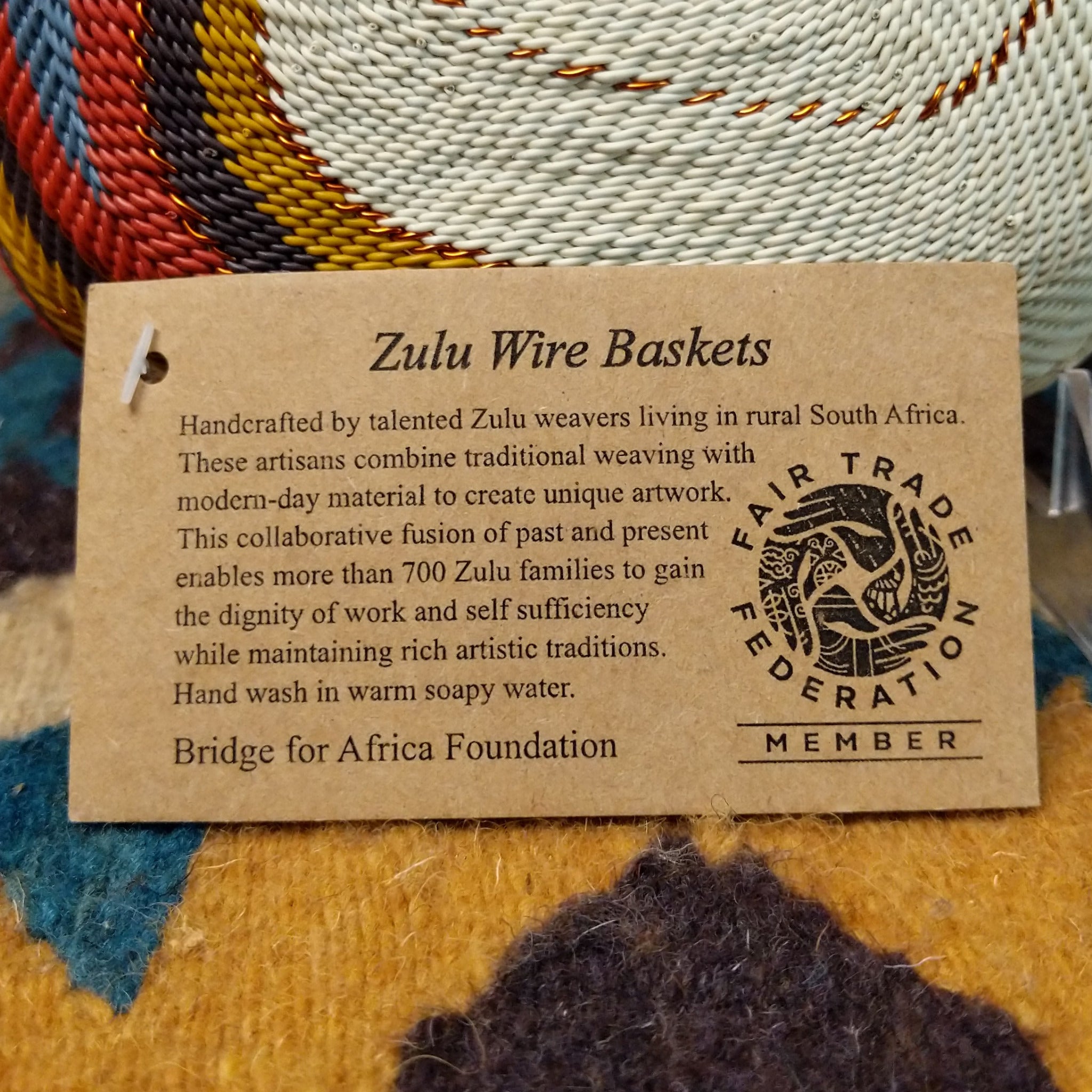 Handwoven square wire art bowl by Bridge for Africa featuring a spiral pattern in recycled wire with ivory, rust, blue, gold, brown, and charcoal tones, ideal for home décor or wall display; artisan information.