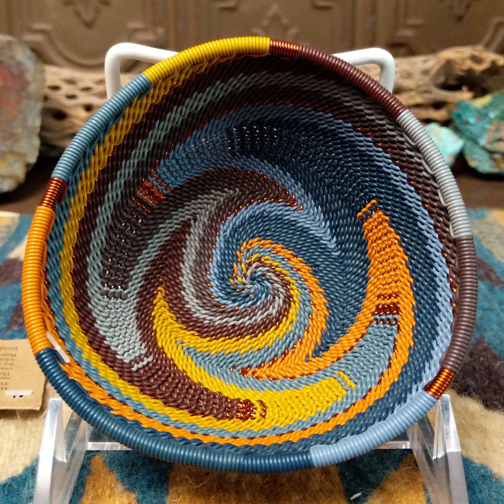 Handwoven triangular wire art bowl by Bridge for Africa featuring a spiral pattern in recycled wire with slate blue, mustard yellow, charcoal, brown, and gray tones, ideal for home décor or wall display.