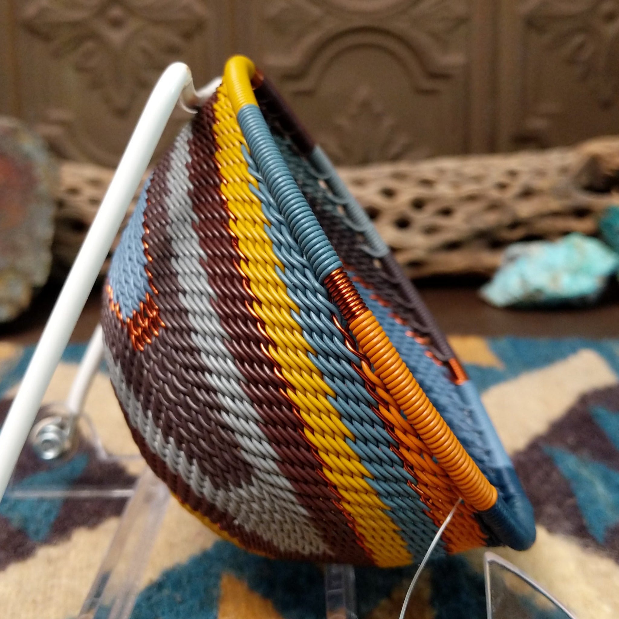 Handwoven triangular wire art bowl by Bridge for Africa featuring a spiral pattern in recycled wire with slate blue, mustard yellow, charcoal, brown, and gray tones, ideal for home décor or wall display; side view.