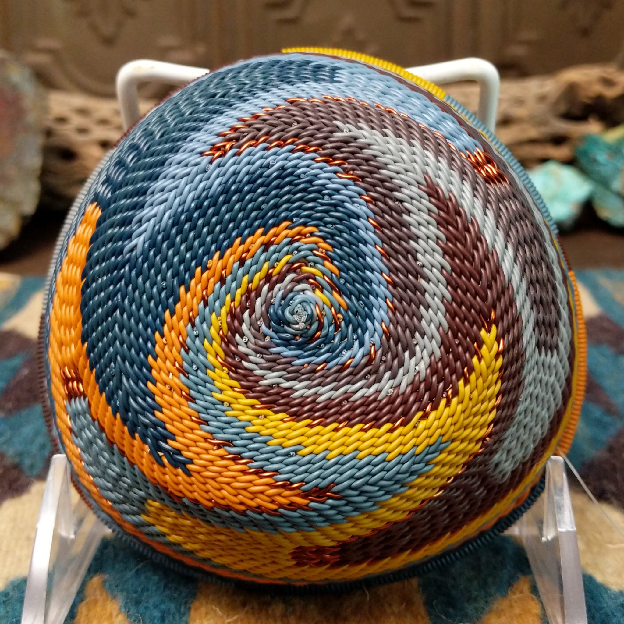 Handwoven triangular wire art bowl by Bridge for Africa featuring a spiral pattern in recycled wire with slate blue, mustard yellow, charcoal, brown, and gray tones, ideal for home décor or wall display; underside view.