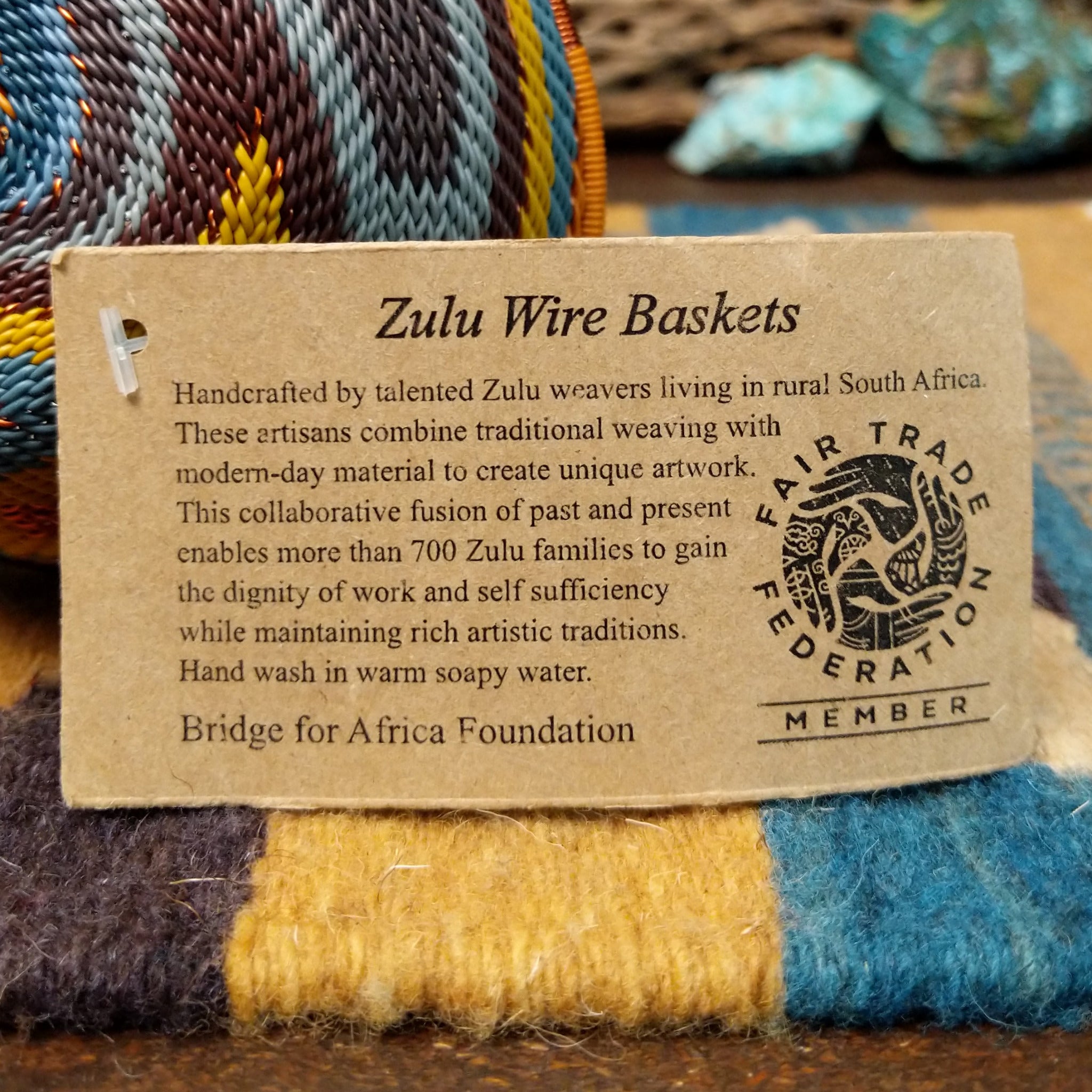 Handwoven triangular wire art bowl by Bridge for Africa featuring a spiral pattern in recycled wire with slate blue, mustard yellow, charcoal, brown, and gray tones, ideal for home décor or wall display; artisan information.