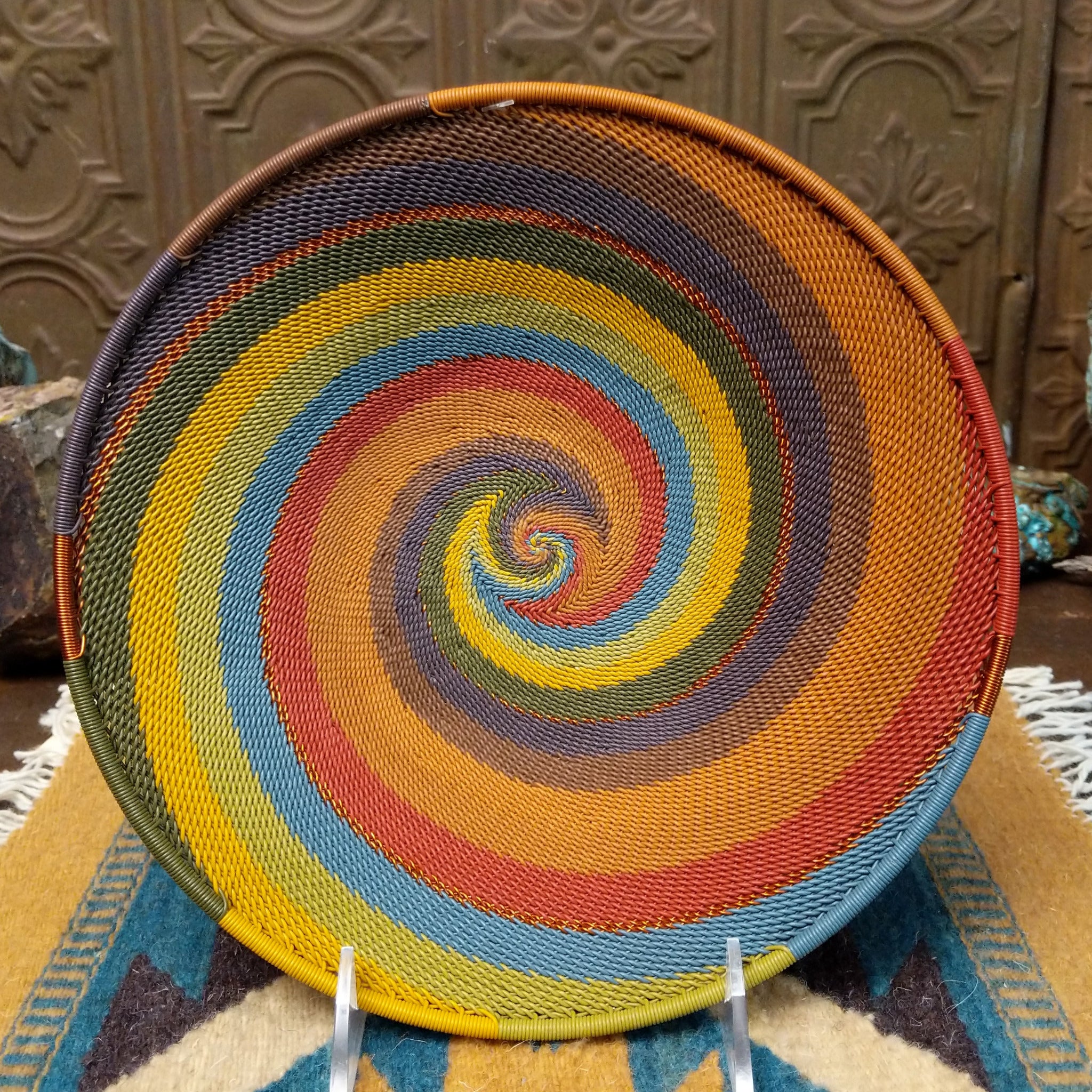 Handwoven wire art bowl by Bridge for Africa featuring a spiral pattern in recycled wire with golden yellow, olive green, dusty blue, rust, tan, brown, and charcoal tones, ideal for home décor or wall display.