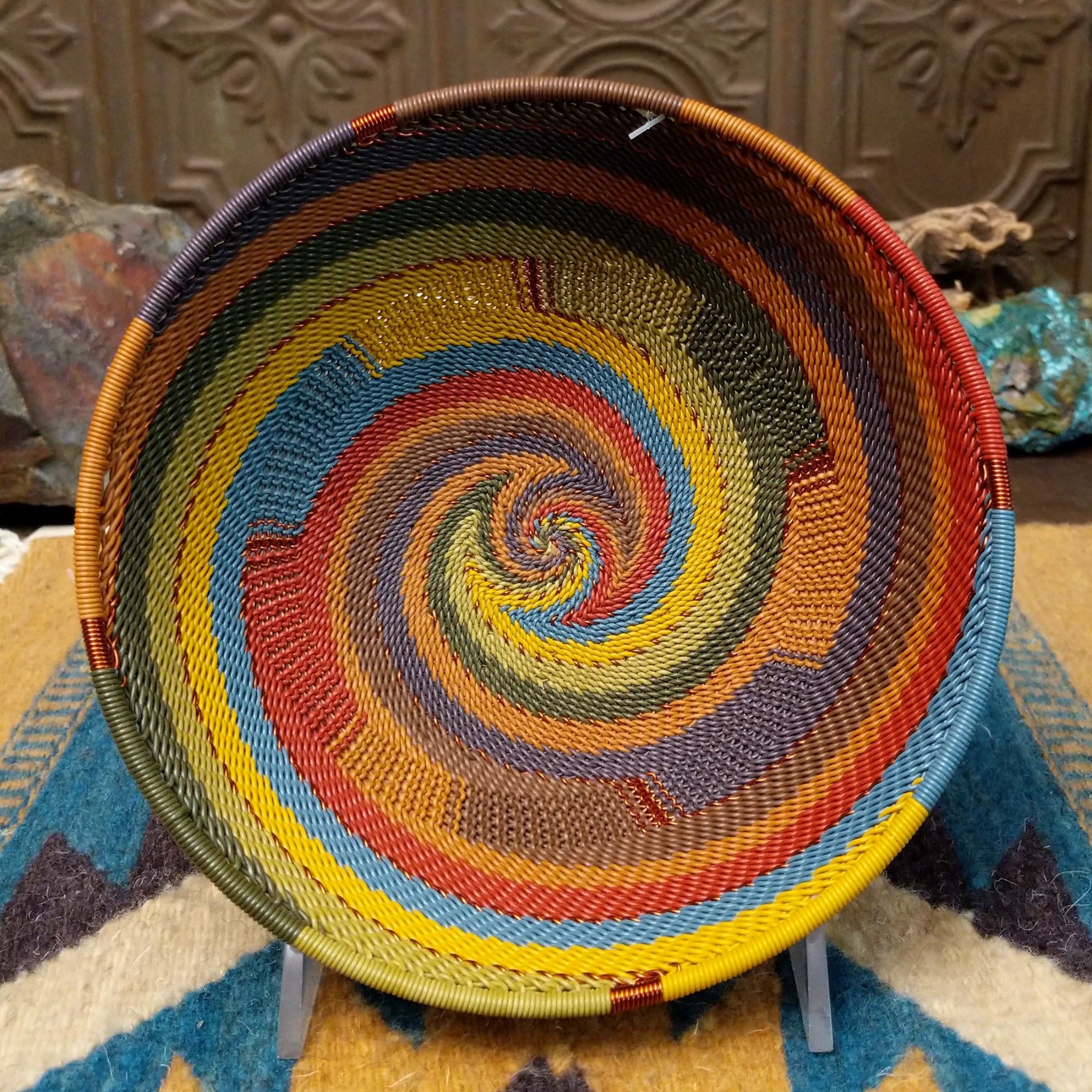 Handwoven wire art bowl by Bridge for Africa featuring a spiral pattern in recycled wire with olive green, mustard yellow, dusty blue, rust, terracotta, brown, and charcoal tones, perfect for home décor or wall display.