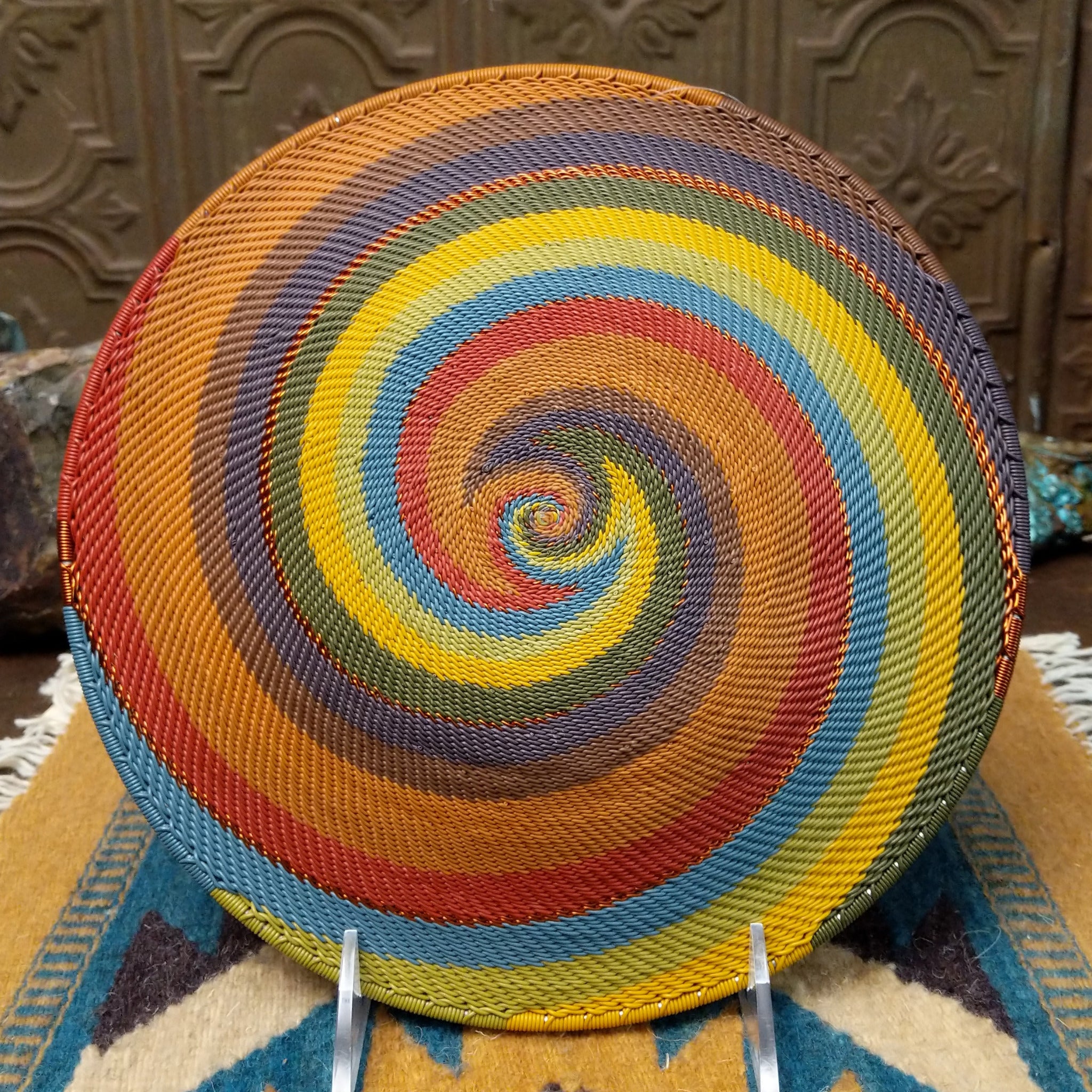 Handwoven wire art bowl by Bridge for Africa featuring a spiral pattern in recycled wire with golden yellow, olive green, dusty blue, rust, tan, brown, and charcoal tones, ideal for home décor or wall display; underside view.