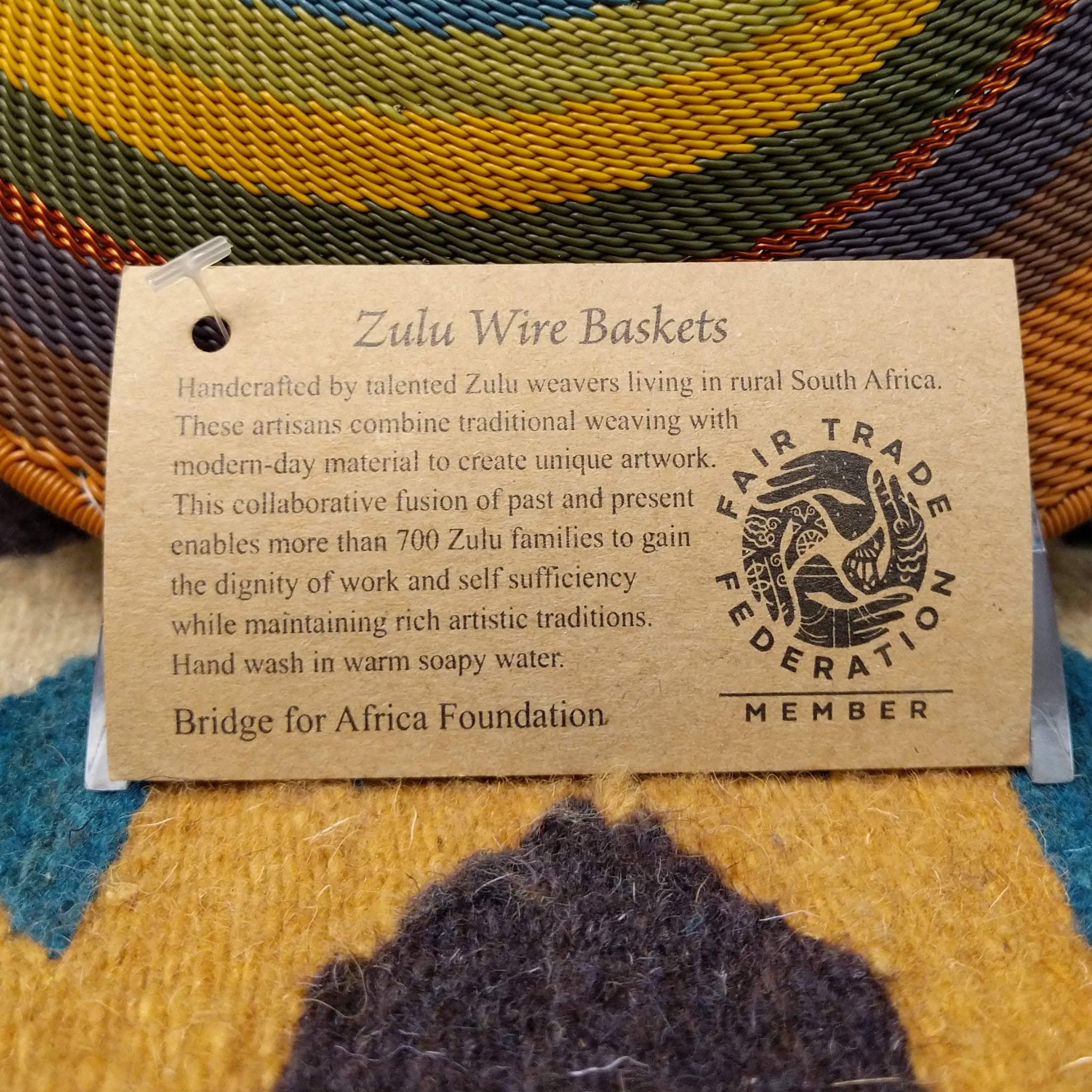 Handwoven wire art bowl by Bridge for Africa featuring a spiral pattern in recycled wire with golden yellow, olive green, dusty blue, rust, tan, brown, and charcoal tones, ideal for home décor or wall display; artisan information.