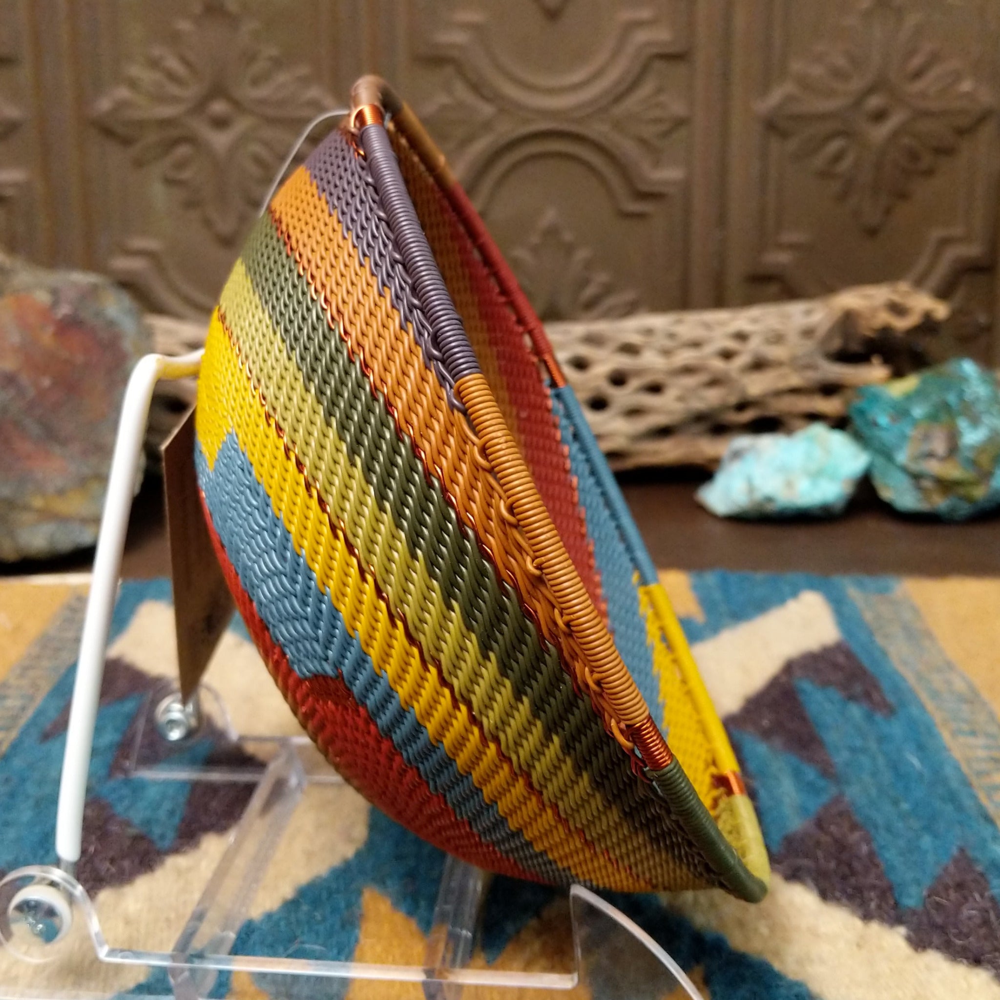 Handwoven wire art bowl by Bridge for Africa featuring a spiral pattern in recycled wire with olive green, mustard yellow, dusty blue, rust, terracotta, brown, and charcoal tones, perfect for home décor or wall display; side view.