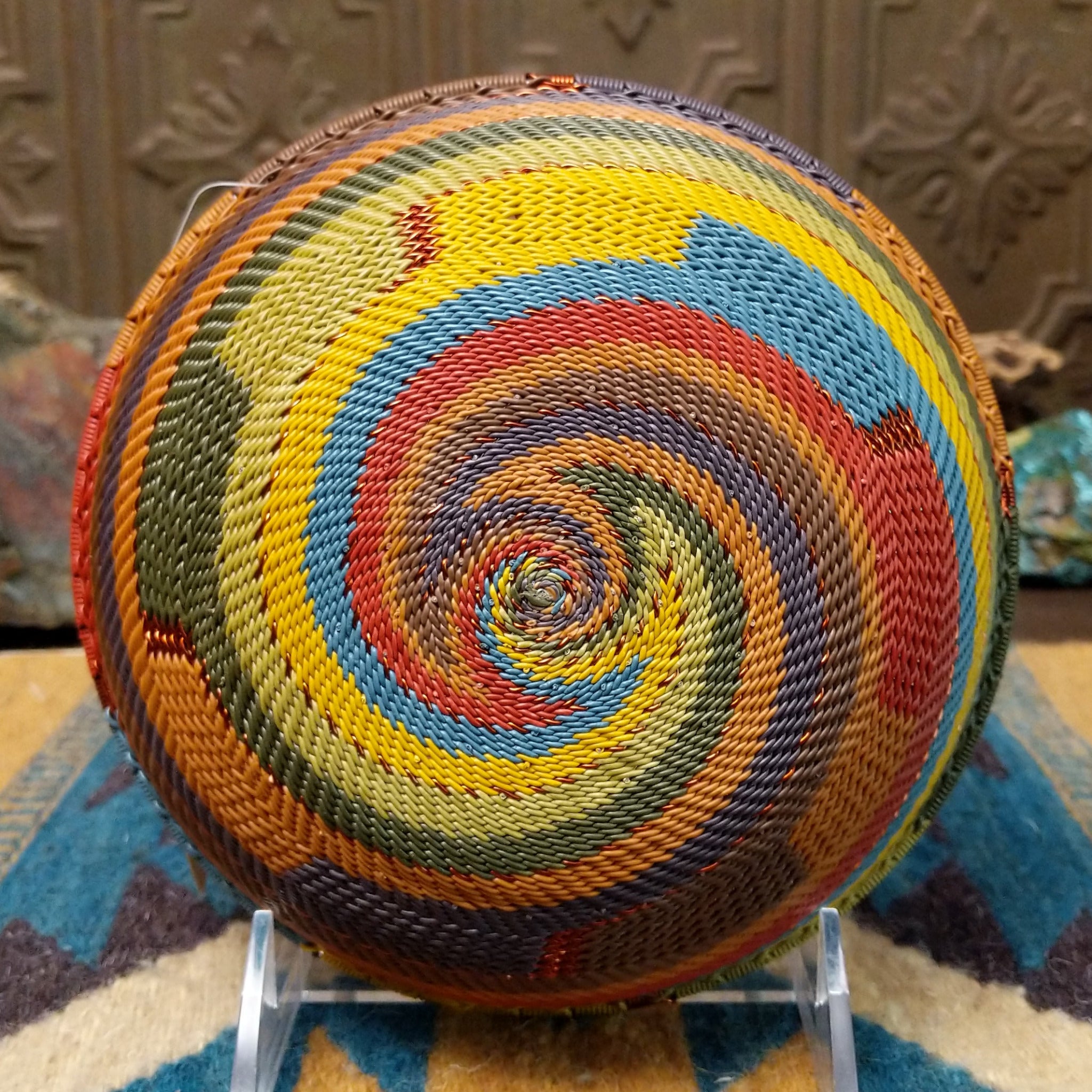 Handwoven wire art bowl by Bridge for Africa featuring a spiral pattern in recycled wire with olive green, mustard yellow, dusty blue, rust, terracotta, brown, and charcoal tones, perfect for home décor or wall display; underside view.