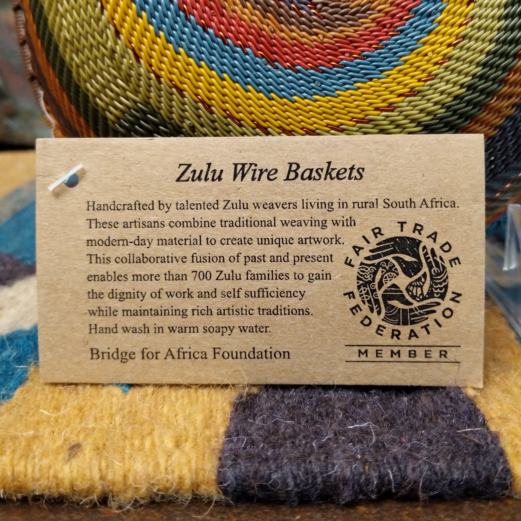 Handwoven wire art bowl by Bridge for Africa featuring a spiral pattern in recycled wire with olive green, mustard yellow, dusty blue, rust, terracotta, brown, and charcoal tones, perfect for home décor or wall display; artisan information.